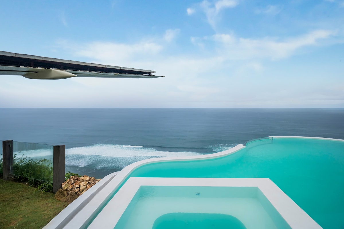An infinity pool extends toward the horizon, blending seamlessly with the ocean. A jacuzzi is nestled beside the pool, offering unobstructed views of the waves, while lush greenery frames the area. The sky reflects soft hues of blue, enhancing the tranquil ambiance.