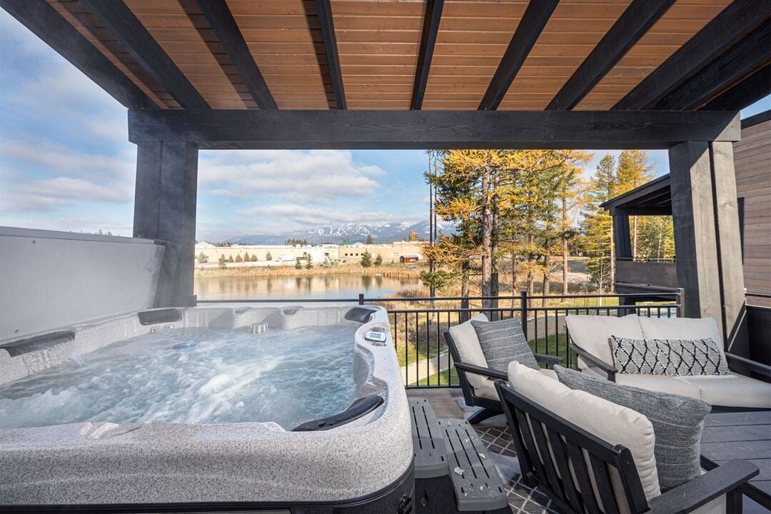 A private hot tub is positioned on a covered patio, providing views of Eagle Lake and surrounding mountains. Comfortable outdoor seating is arranged nearby, with soft cushions inviting relaxation. The backdrop features a mix of trees and distant snow-capped peaks under a clear sky.