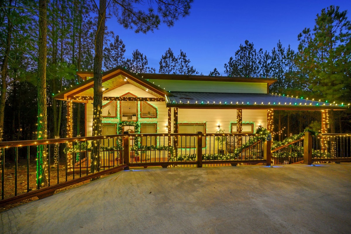 The cabin's exterior is adorned with festive lighting, highlighting its architectural features against a twilight backdrop. Surrounded by tall pine trees, the inviting space is accessible via a ramp, enhancing its welcoming ambiance in a serene forest setting.