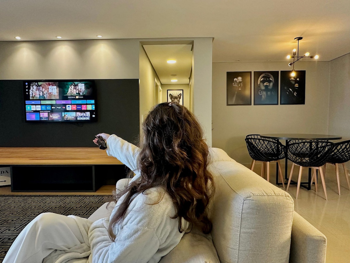 A modern living area features a neutral-colored couch in the foreground, with a large wall-mounted television displaying a streaming interface. A dining table with stylish black chairs is visible in the background. Soft lighting creates a cozy atmosphere.