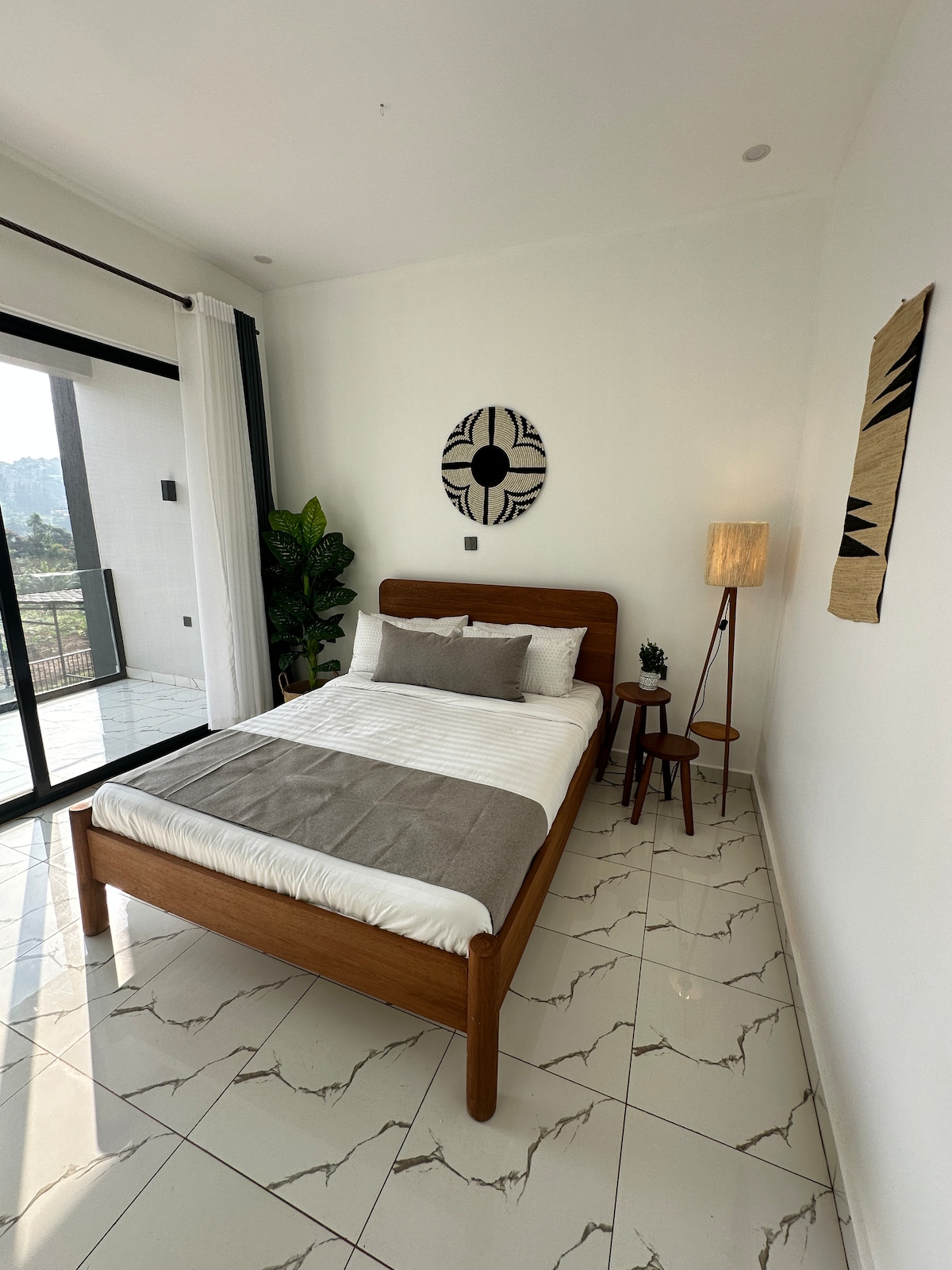 A well-appointed room features a double bed with neutral linens laid over a light wooden frame. A tall lamp stands next to a small side table, while a decorative wall hanging and a plant add character to the space. Large glass doors reveal views of the outdoors.