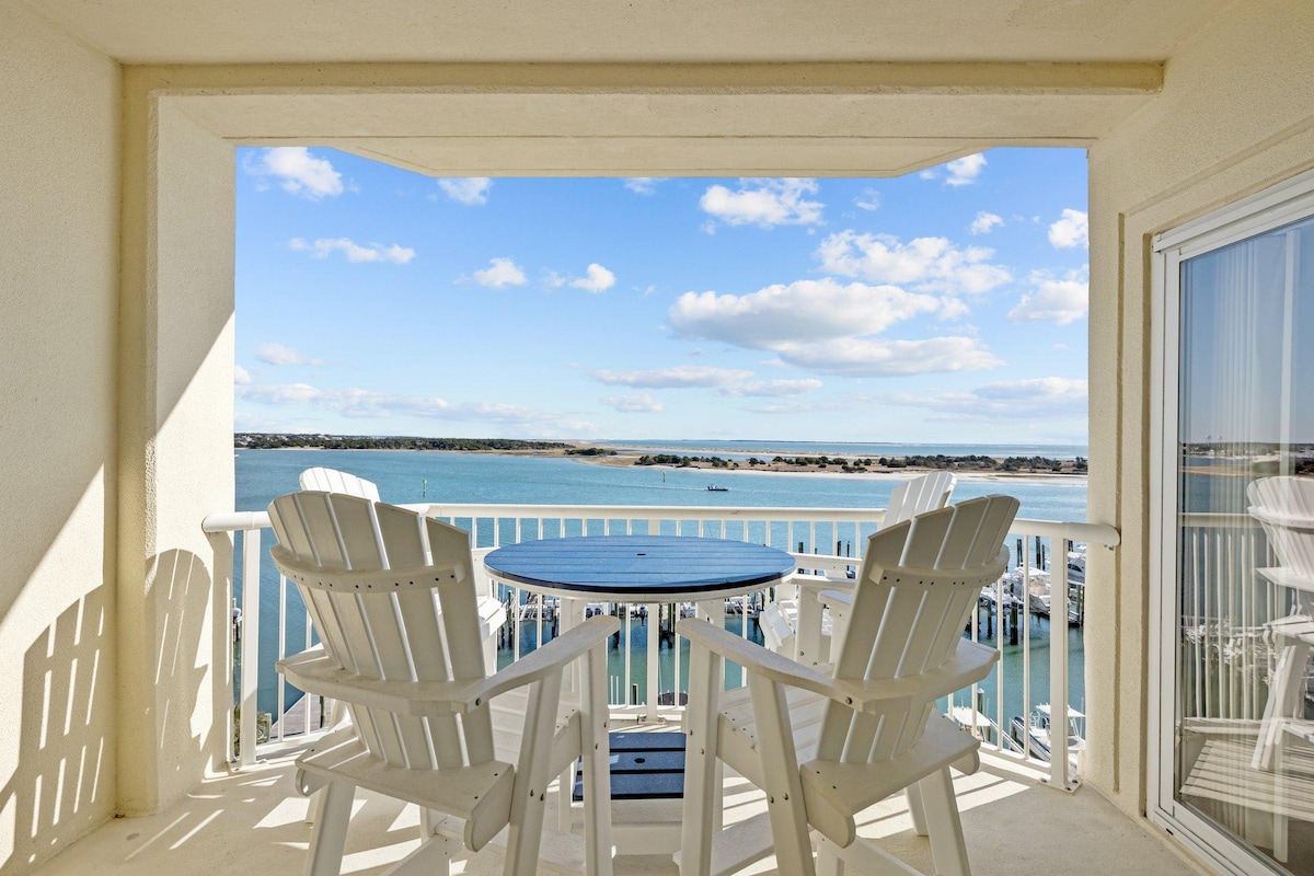 A covered balcony offers a round table with two adirondack chairs, overlooking the water. The view includes a marina with boat slips and serene blue waters under a bright sky, providing a peaceful outdoor space for relaxation.