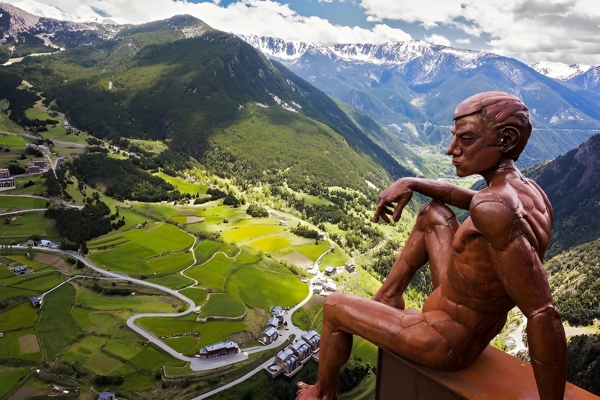 A sculpture of a seated figure overlooks a mountainous landscape, surrounded by lush greenery and winding roads. The backdrop features snow-capped peaks that merge into a cloudy blue sky, creating a serene and expansive view of the valley below.