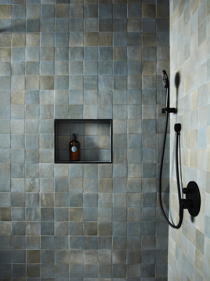 The shower space features a textured tile wall in varying shades of blue and gray, complemented by a sleek black shower fixture. A built-in shelf holds a small bottle, adding to the modern aesthetic of the space.