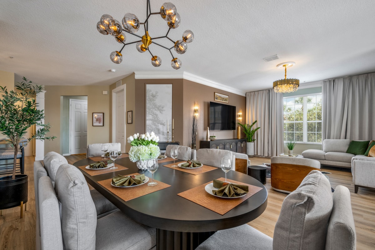 The dining area features a large oval table surrounded by eight comfortable, cushioned chairs. Soft lighting from a distinctive chandelier adds warmth to the space, while contemporary decor elements create a modern atmosphere. A nearby lounge area includes a plush sofa, inviting relaxation.
