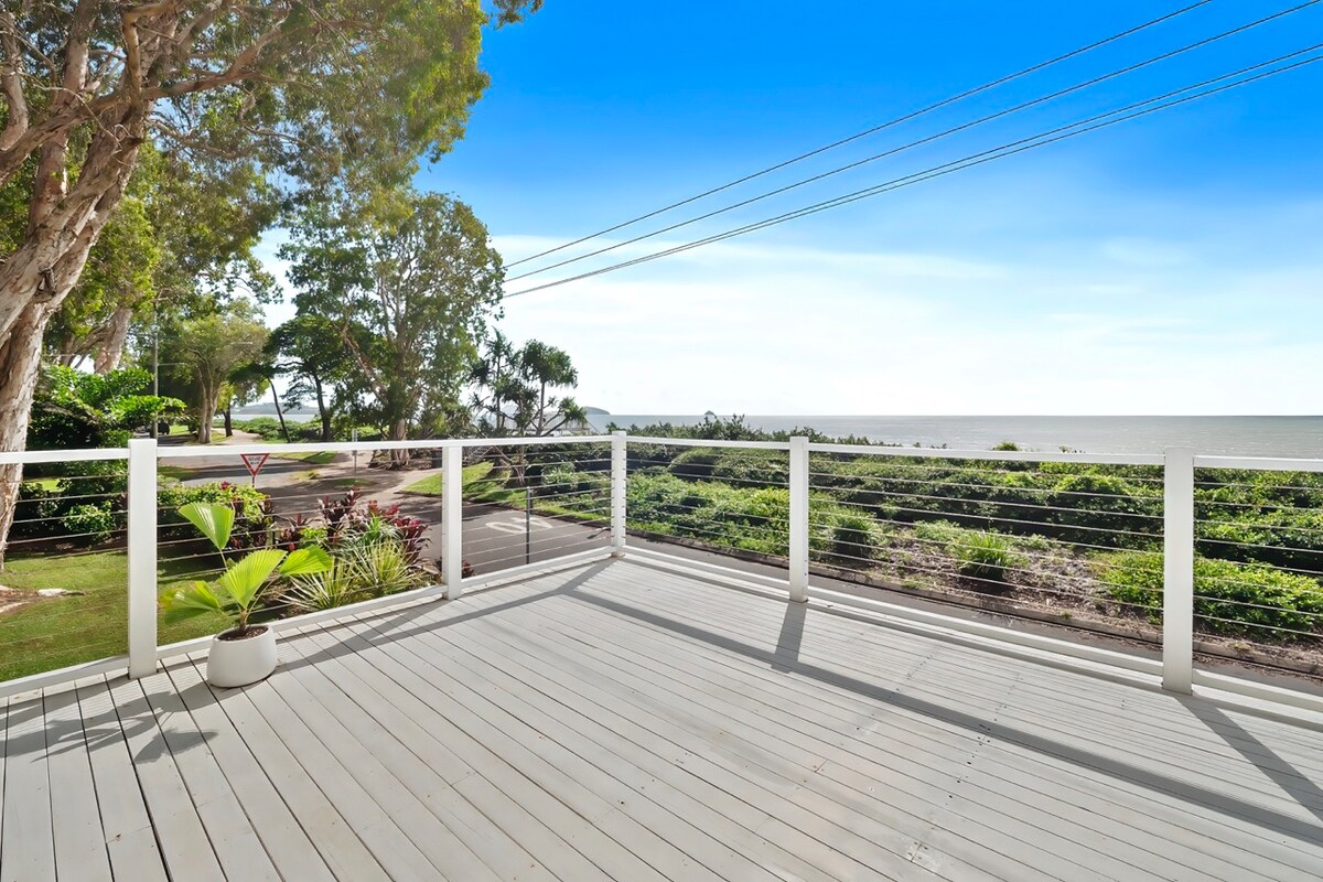 A spacious deck features a clean, wooden surface and a white railing, providing unobstructed views of the ocean. Lush greenery surrounds the area, and a potted plant adds a touch of nature. The tranquil ocean can be seen in the distance under a clear blue sky.