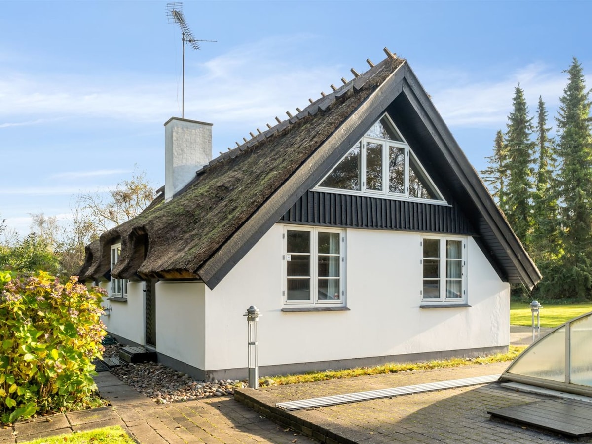 The exterior of a charming single-family house is shown, featuring a thatched roof and a combination of white and black siding. Large windows provide ample natural light, while neatly landscaped gardens surround the property, enhancing its serene setting.