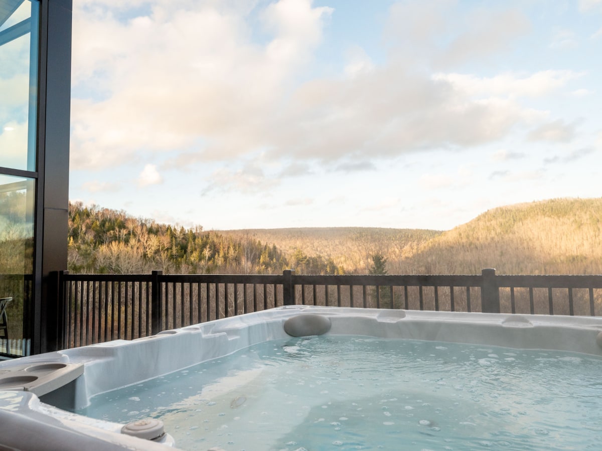 The private deck features a hot tub filled with water, overlooking a valley bordered by lush green mountains. The sky above is partly cloudy, allowing natural light to illuminate the serene outdoor space.