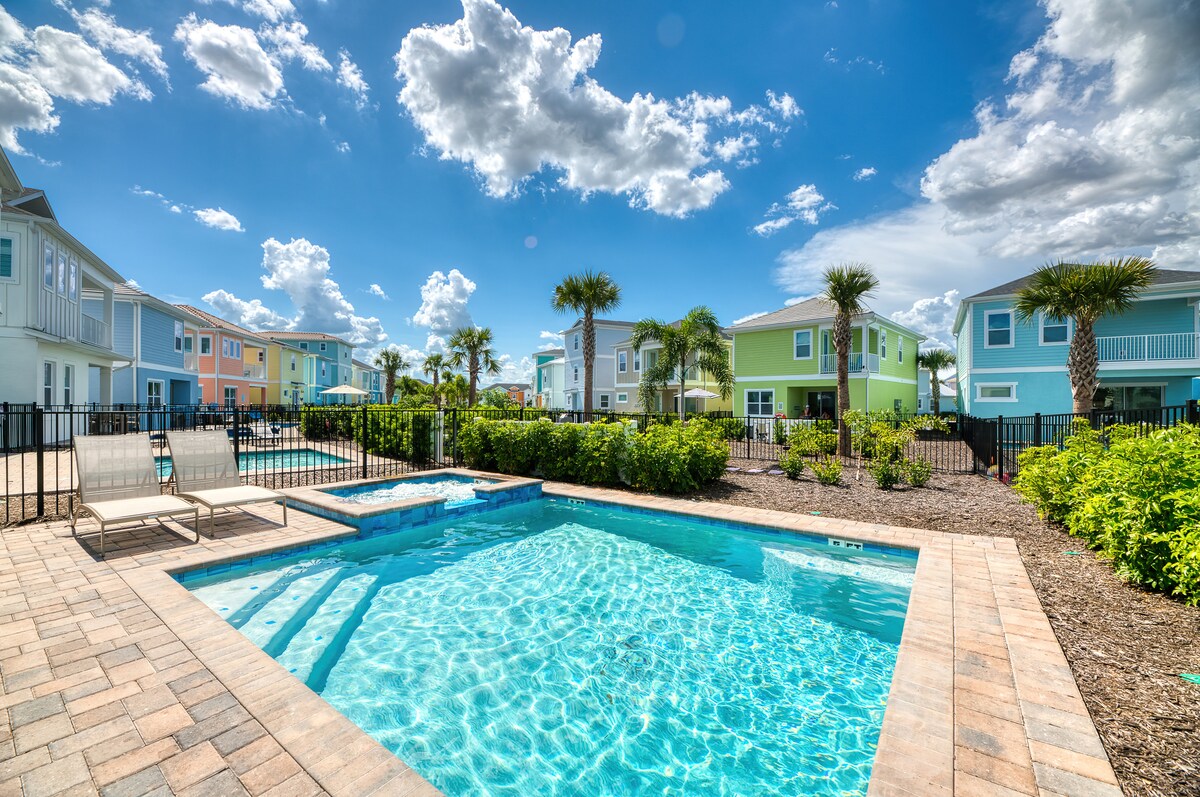 A refreshing pool is surrounded by landscaped grounds and colorful cottages. Palm trees add a tropical feel, while a hot tub is located beside the pool. A paved area with lounge chairs is visible, inviting relaxation under the sun.