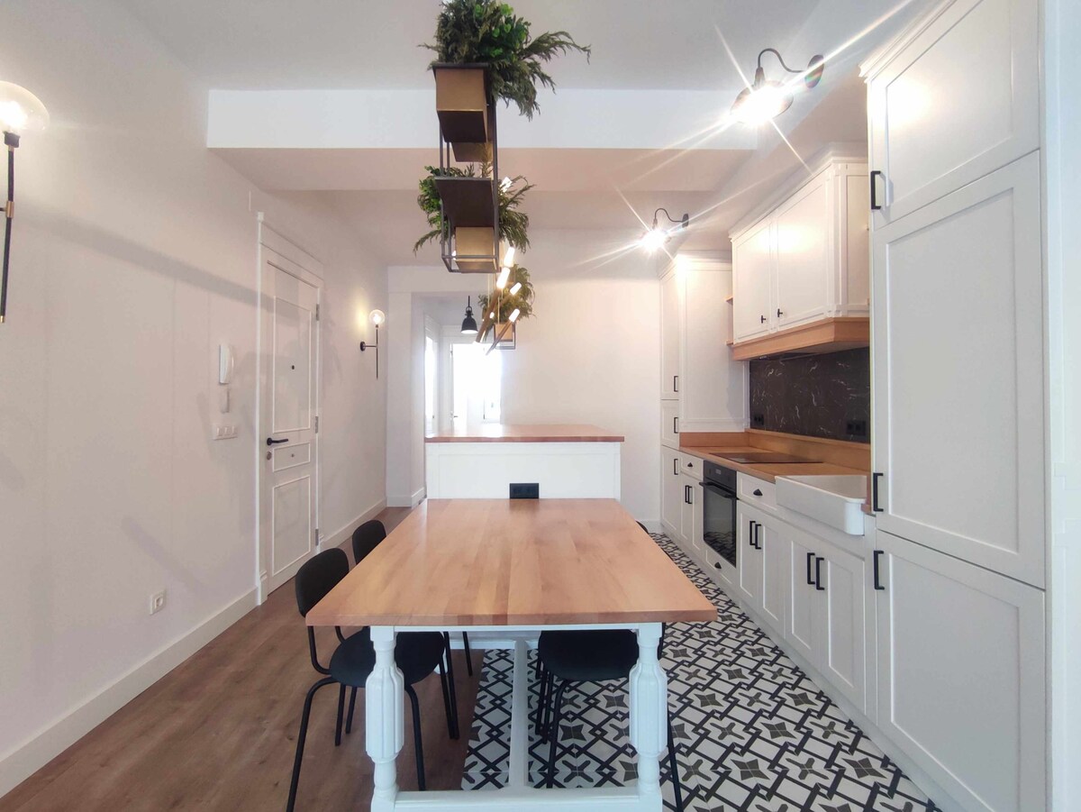 The kitchen area is characterized by modern cabinetry and a large wooden dining table surrounded by black chairs. Pendant lighting is hung above the table, and decorative plants are displayed from an overhead installation. A patterned floor adds visual interest to the space.