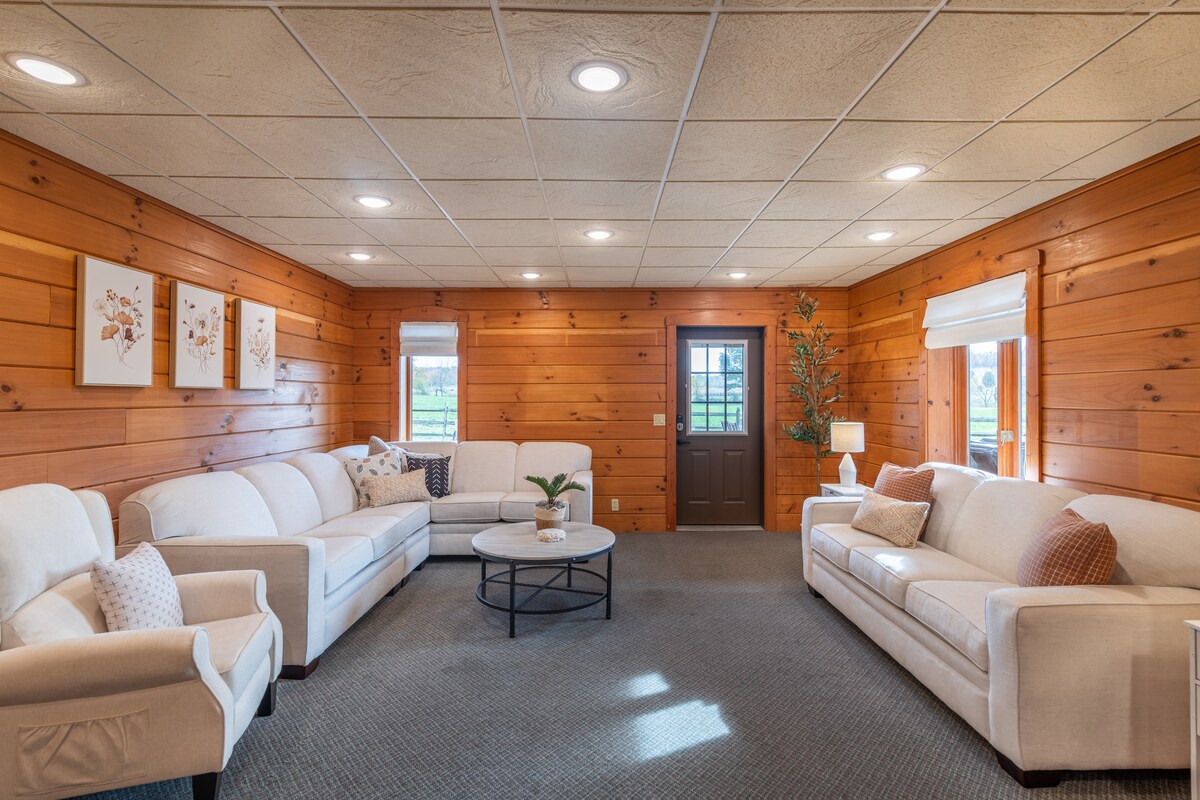 The spacious living area features wooden walls, creating a warm environment. Two comfortable sofas and a pair of armchairs encircle a central coffee table. Natural light enters through a door and windows, enhancing the cozy seating arrangement.