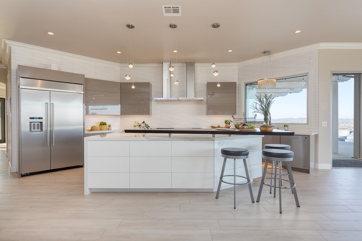A modern chef's kitchen features sleek cabinetry and high-end stainless steel appliances. An island with barstool seating invites gatherings, while large windows provide natural light and views of the surrounding landscape. Minimalist decor and a neutral color palette enhance the inviting ambiance.