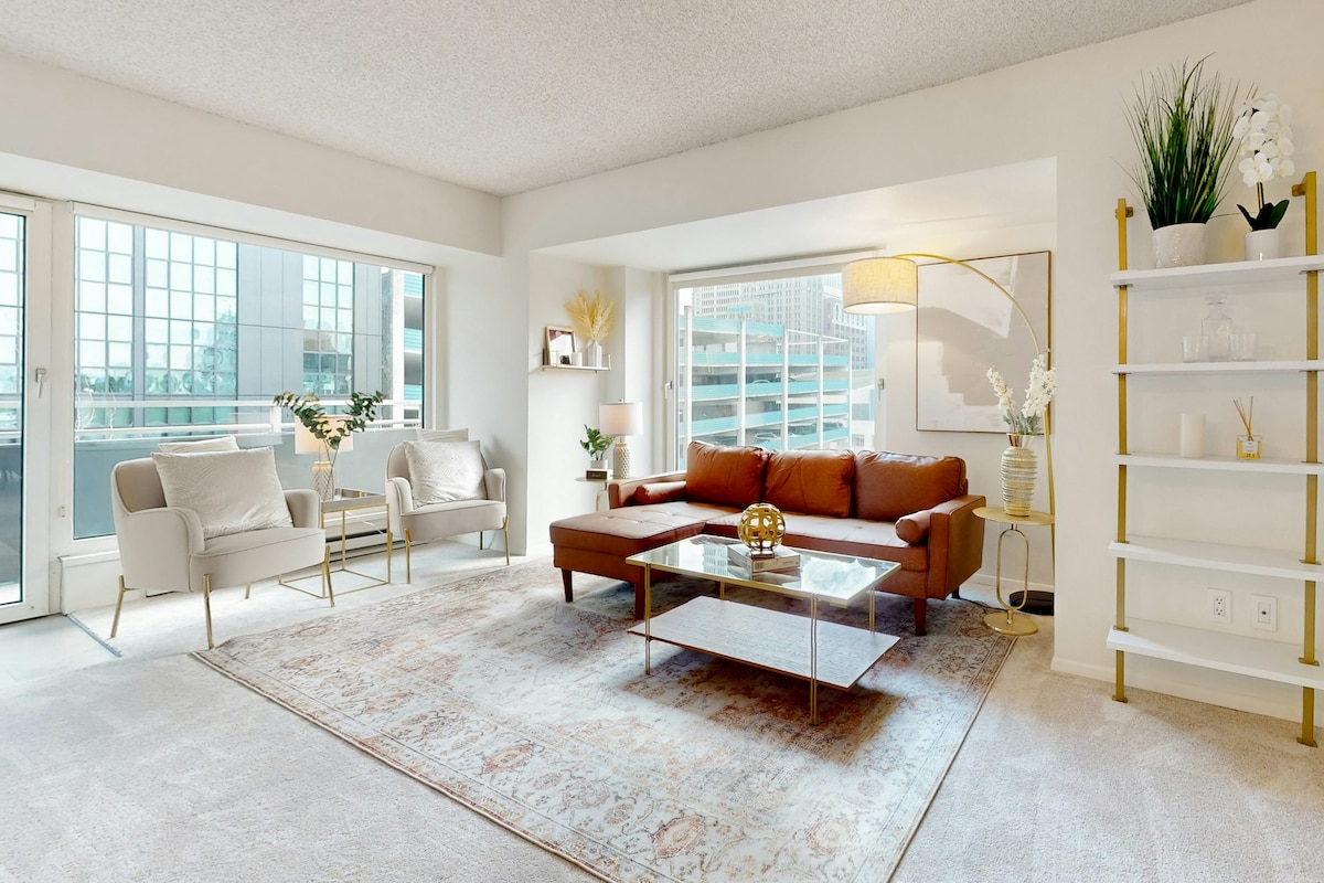 A modern living area features a brown sectional sofa and two light-colored armchairs situated on a soft area rug. Natural light fills the space through large windows, and decorative elements like a vase and artwork enhance the elegant atmosphere.