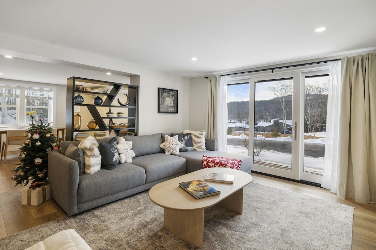 A modern living area is presented with a light gray sectional sofa adorned with decorative pillows. A round coffee table is centrally positioned, and large sliding glass doors lead to an outdoor view, allowing natural light to fill the space.