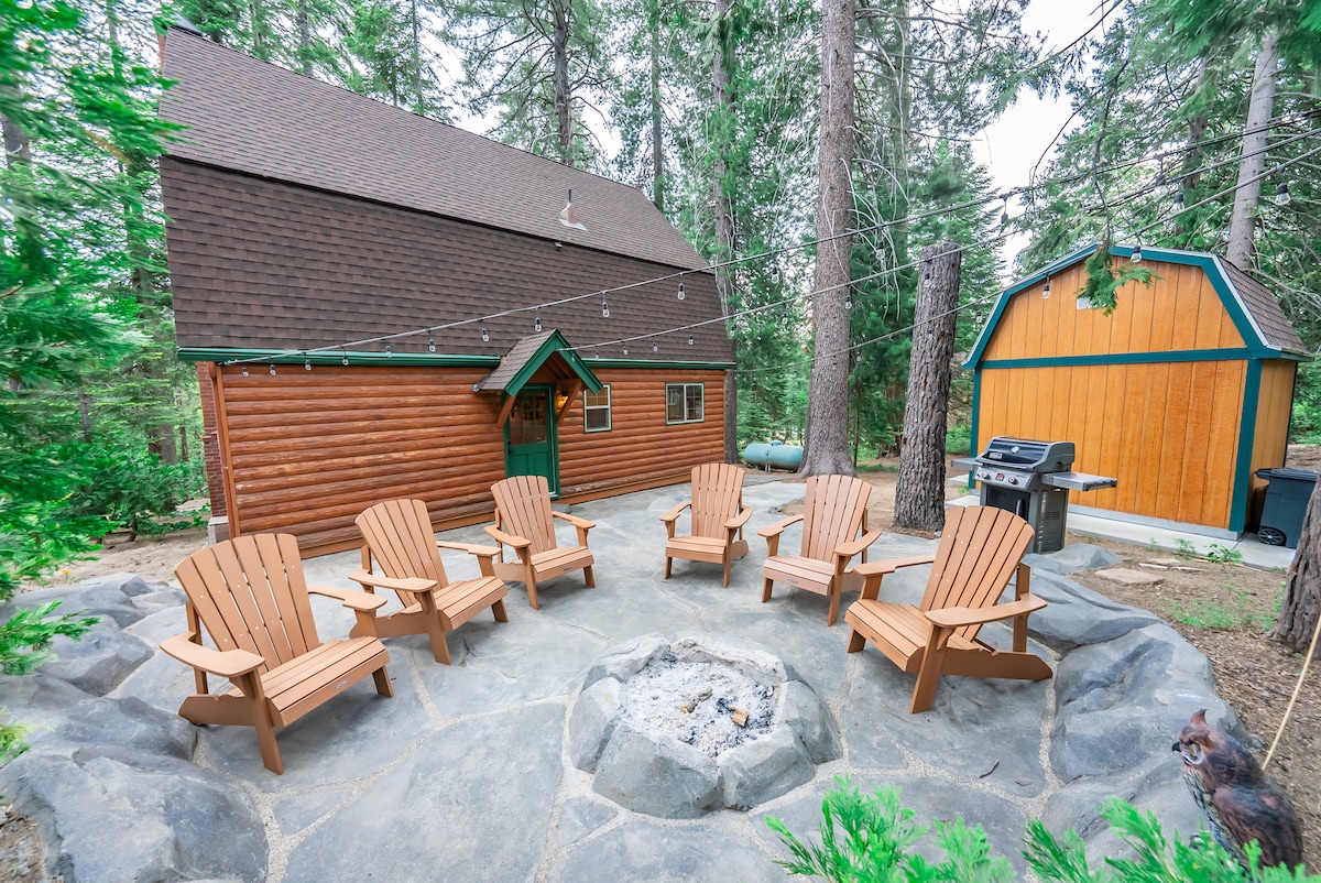 An outdoor gathering area is showcased with six wooden Adirondack chairs arranged in a circular pattern around a stone fire pit. The background highlights a cabin with a brown exterior, surrounded by tall trees, and a shed is visible on the side.