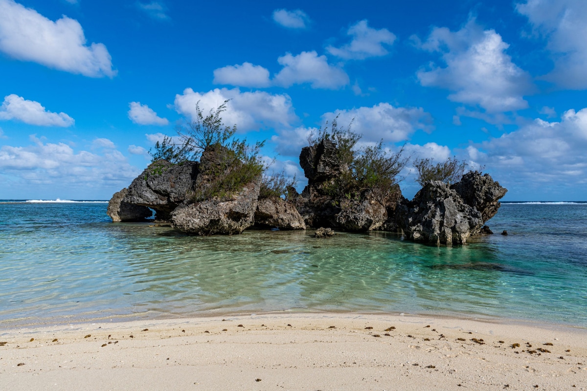 A rugged rock formation rises from the crystal-clear water, surrounded by soft sandy beaches. Lush vegetation clings to the rocks, contrasting against the serene blue sky dotted with fluffy clouds. Gentle waves lap at the shoreline, creating a peaceful coastal scene.