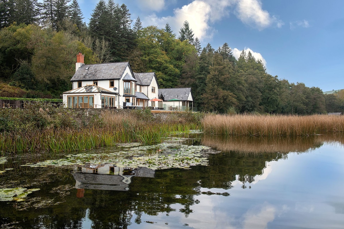 The image depicts a charming countryside house set against a backdrop of lush trees, reflecting beautifully in the calm waters of a lake. Reeds and lily pads are visible in the foreground, enhancing the tranquil atmosphere of the picturesque setting.