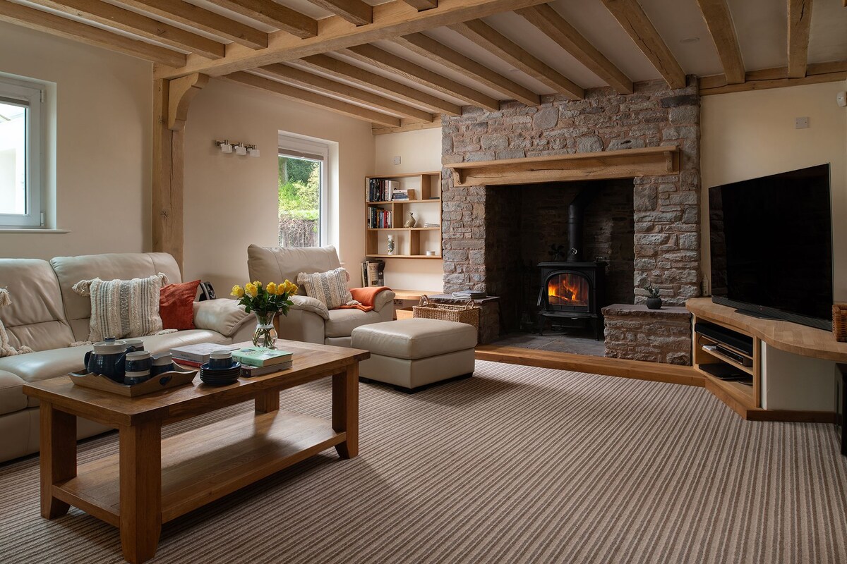 A spacious lounge features exposed wooden beams and a stone fireplace with a glowing fire. Comfortable seating includes a large sectional sofa and two armchairs. A coffee table stands centrally, surrounded by warm tones and soft textures, creating a welcoming environment for relaxation.