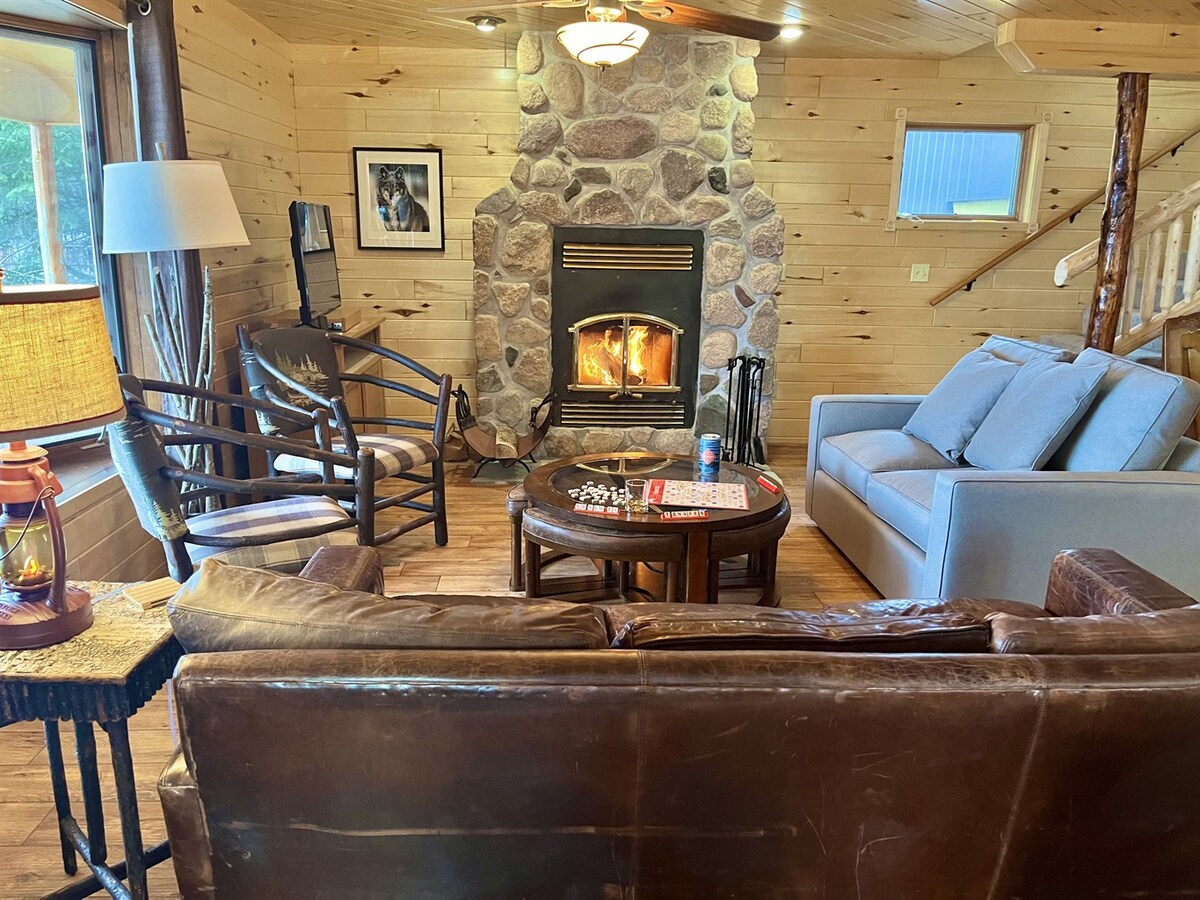 A rustic living room features a stone fireplace as the focal point, with a wood-burning fire gently glowing. Comfortable seating is provided by a leather couch and two chairs. Natural light filters through large windows, enhancing the warm wooden interior.