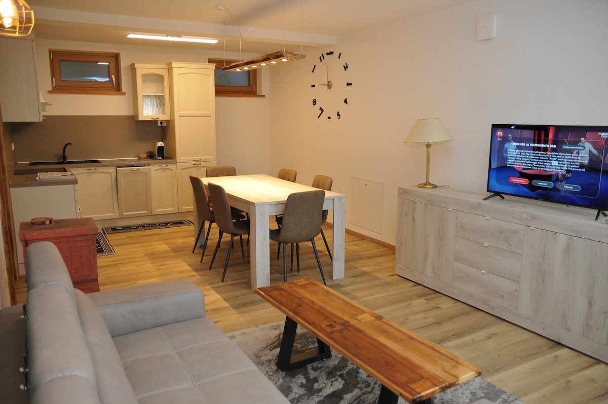 An open-plan living space features a dining area with a large table surrounded by six chairs. A gray sofa is positioned nearby, with a wooden coffee table in front. A modern kitchen is visible, equipped with light cabinetry and appliances. Natural light enters through two windows.