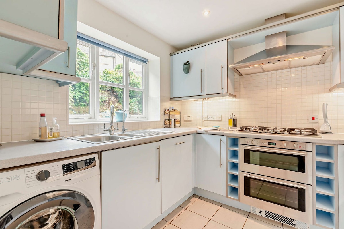 The kitchen features light blue cabinetry and a window allowing natural light. Equipped with an electric oven, gas hob, dishwasher, and washing machine, it provides ample counter space for meal preparation. Shelves are visible, showcasing a selection of cooking essentials.