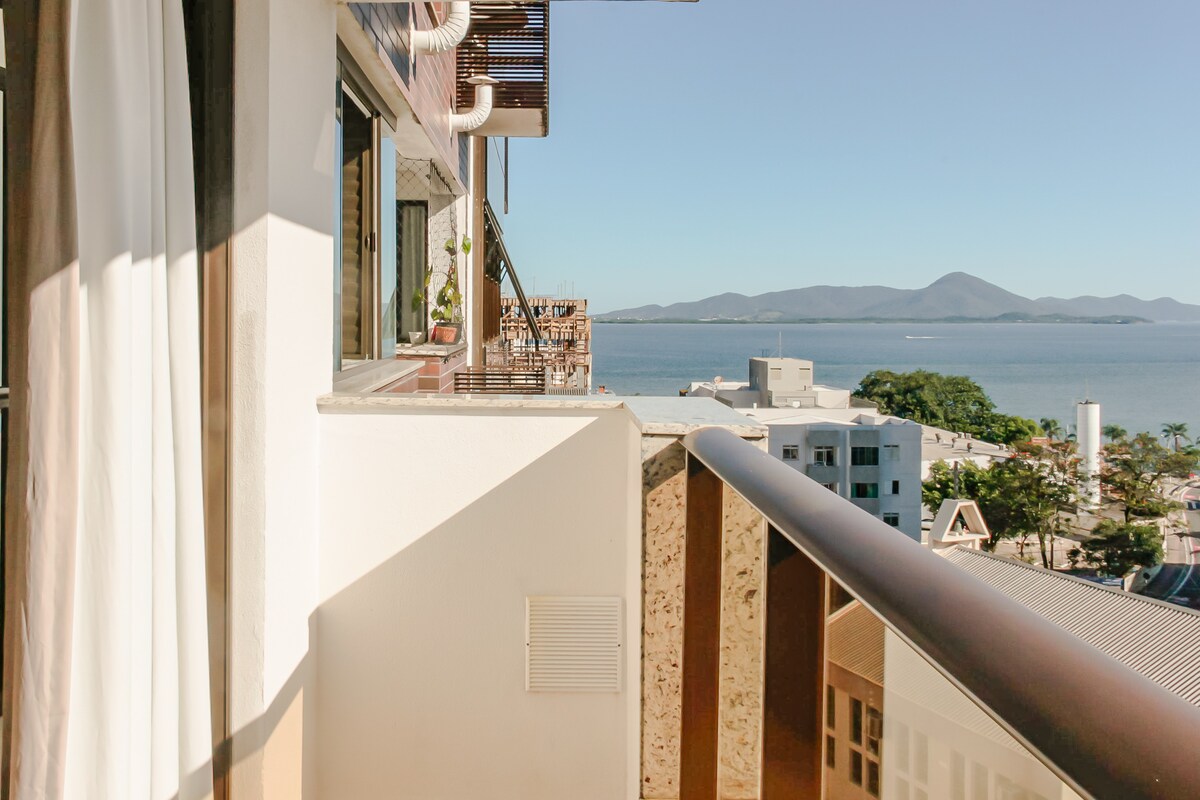 A balcony offers a broad view of the continental bay, with distant mountains visible against a clear blue sky. The space is framed by a sturdy railing, and potted plants add a touch of greenery to the area.