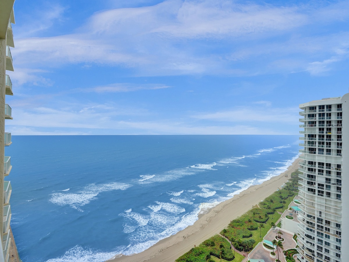 An expansive ocean view is presented from the 26th floor, showcasing rolling waves meeting a sandy shore. The horizon is lined with clear blue skies, while coastal greenery is subtly visible along the beach. The scene captures a sense of tranquility and openness.