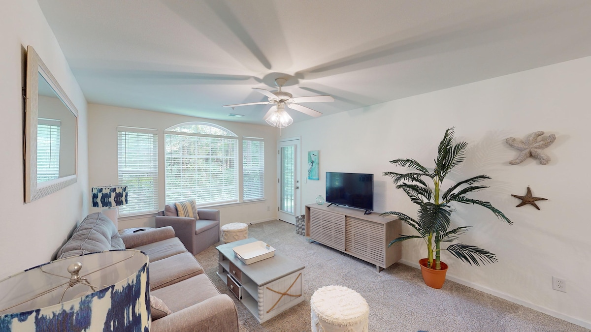 A spacious living area features large windows providing natural light. A sectional sofa, a television on a media console, and decorative elements like a potted plant are visible. Light-colored walls complement the soft carpet, creating a comfortable space for relaxation.
