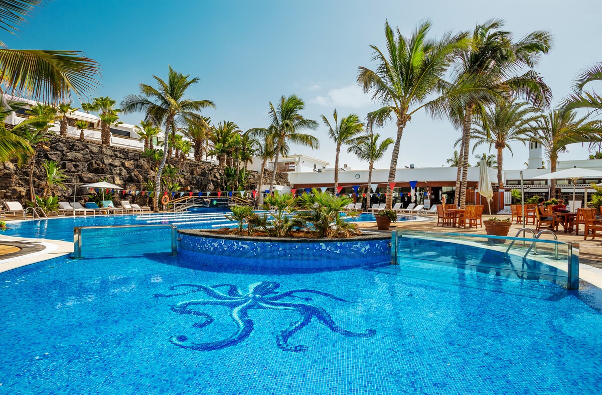 A large outdoor pool area is surrounded by palm trees and lush greenery. The pool features intricate tile patterns in vibrant blue water. Sun loungers are arranged around the pool, providing ample relaxation space for guests. Facilities and seating areas are visible in the background.