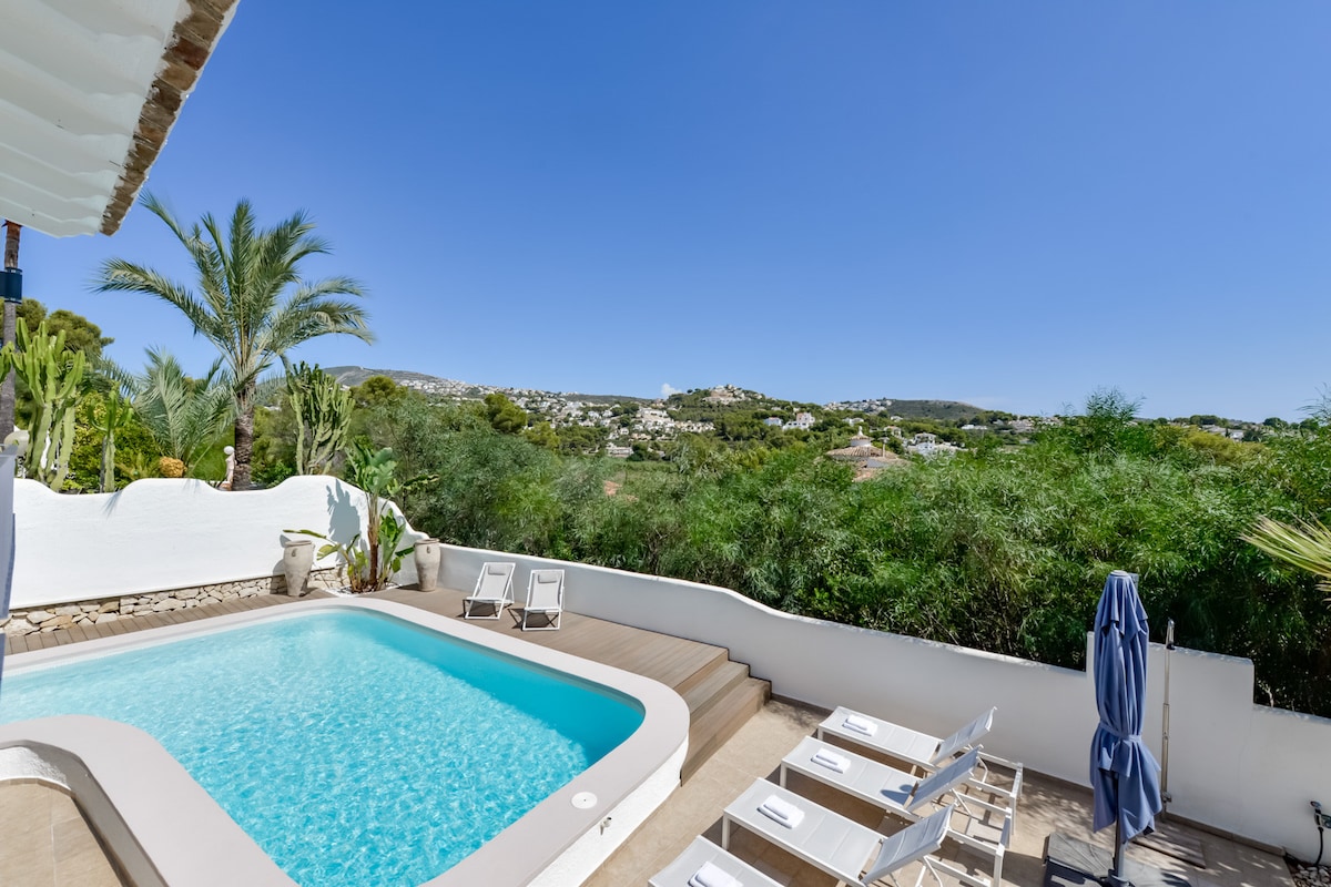 A private swimming pool is framed by a spacious terrace, creating an inviting outdoor area. Surrounding greenery and palm trees enhance the tranquil setting, while distant views of the hills and nearby community can be seen under a clear blue sky.