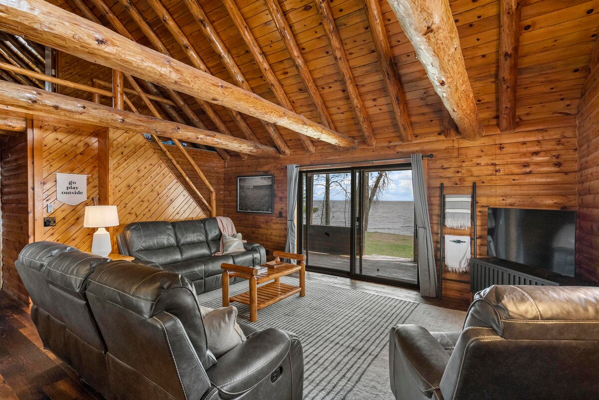 A welcoming living area features vaulted wooden ceilings and large log beams. Comfortable leather recliners are arranged around a coffee table, while a rug adds warmth. Sliding glass doors provide a view of the lake, enhancing the connection to nature.