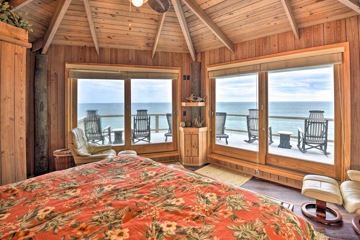 A spacious bedroom features a king bed adorned with a floral patterned blanket. The wood-paneled walls and ceiling provide a warm atmosphere. Large windows frame a view of the ocean and a patio with Adirondack lounge chairs. Natural light fills the room, enhancing the tranquil setting.