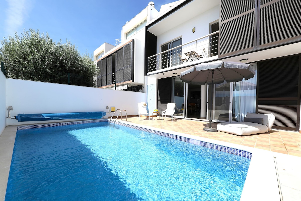 A private pool is surrounded by tiled flooring, with a comfortable sunbed and an umbrella positioned nearby for shade. The villa features large glass doors leading to modern indoor spaces, enhancing connectivity with outdoor leisure areas.