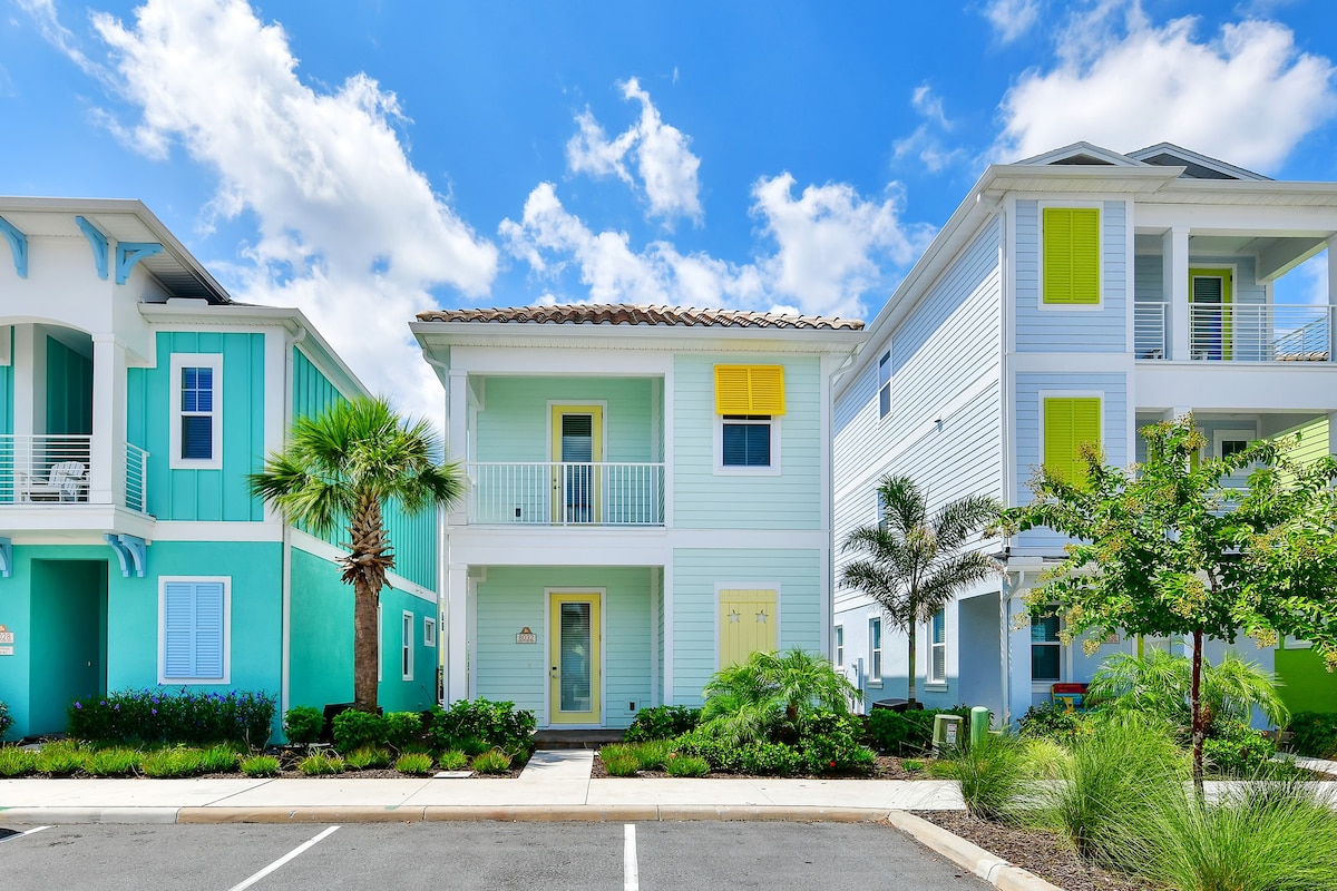 A row of colorful cottages is set against a bright blue sky, featuring palm trees and lush landscaping in the foreground. The cottage exteriors exhibit vibrant colors, with yellow shutters and a welcoming entrance. The inviting atmosphere highlights the resort's charming architecture.