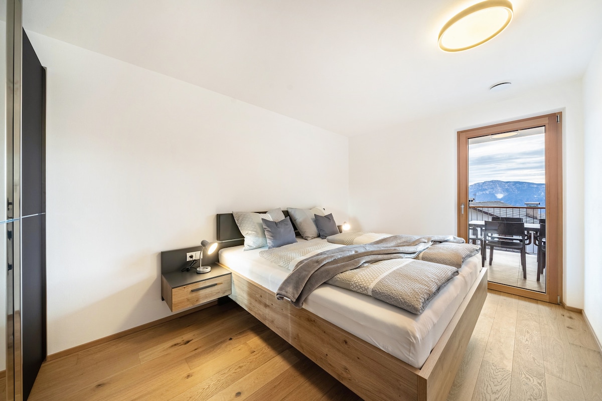 A spacious bedroom is displayed, featuring a large bed covered with soft bedding and coordinated pillows. Nightstands are positioned on either side, each equipped with a lamp. A glass door provides access to a private terrace, while natural light brightens the room.