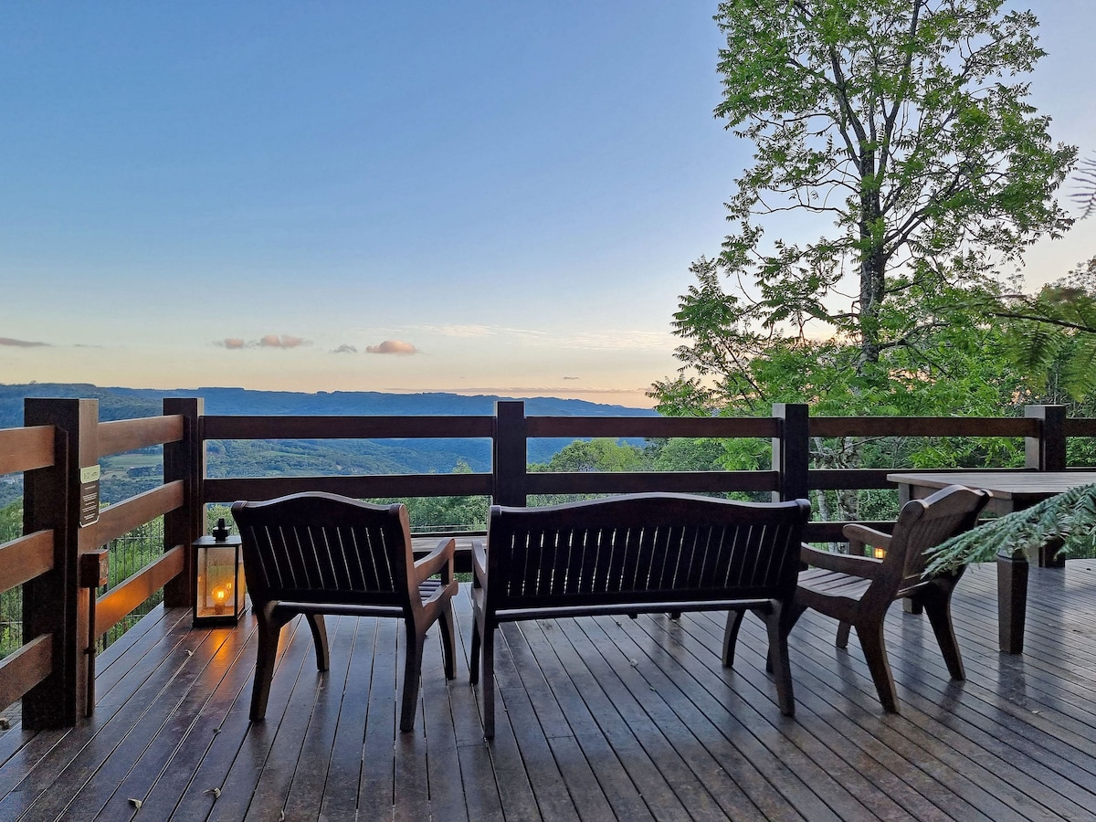A wooden deck is outfitted with comfortable outdoor seating, offering a peaceful view of the valley and distant hills. A lantern adds a subtle touch of warmth, while lush greenery surrounds the area, enhancing the connection to nature.
