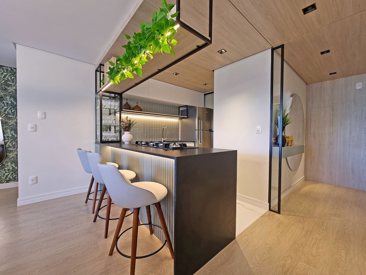 A modern kitchen is presented with an island that includes seating for three. Greenery is displayed above the counter, while sleek cabinetry and stainless appliances provide a contemporary feel. Soft lighting enhances the open space, contributing to a welcoming cooking environment.