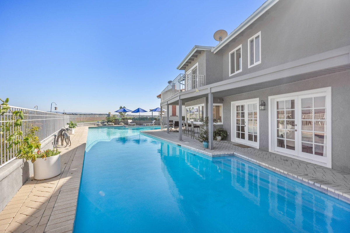 A tranquil outdoor area features a large swimming pool surrounded by a spacious deck. The pool's clear water extends towards a nearby patio area, furnished with seating and tables. Bright blue umbrellas provide shaded spots, and the home showcases large windows that enhance natural light.