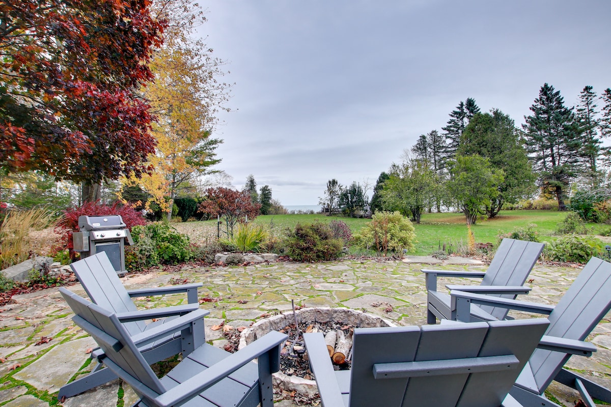 A stone patio features a fire pit surrounded by four Adirondack chairs, inviting relaxation outdoors. Lush greenery and trees are visible in the background, along with a glimpse of Lake Michigan's tranquil waters, creating a serene outdoor space for gatherings.