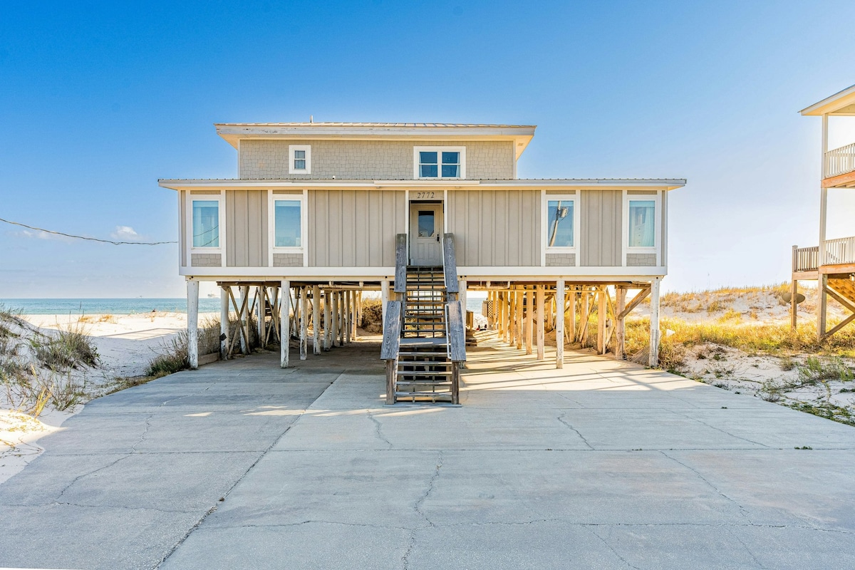 The beach house is elevated on stilts, featuring a wide stairway leading to the main entrance. Large windows frame the upper deck, offering views of the sandy beach and the ocean, while a concrete area beneath provides ample parking.