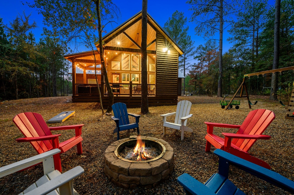 An outdoor seating area is arranged around a stone fire pit, featuring brightly colored chairs in red, blue, and white. The cabin is seen in the background, illuminated softly by warm lights, surrounded by trees in a serene setting.