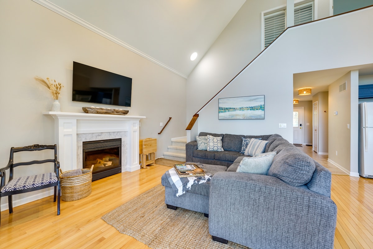 A spacious living area features a comfortable gray sectional sofa, positioned near a gas fireplace below a large wall-mounted television. Natural light enters through high windows, illuminating the warm wooden floors and simple decor. A staircase leads to the upper level, enhancing the open feel.