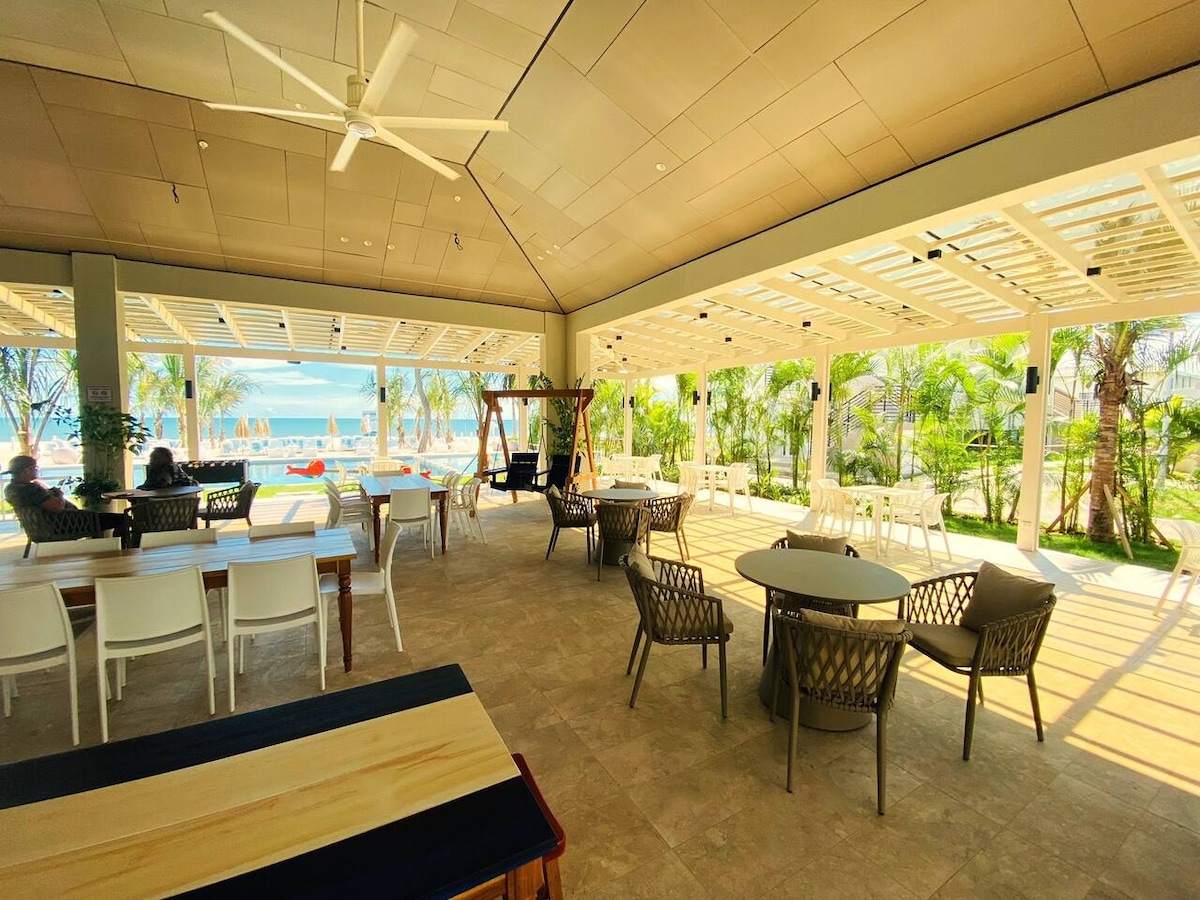 An airy communal dining area features a mix of tables and chairs, with large windows allowing natural light and views of the ocean. Comfortable seating arrangements are positioned around the space, complemented by tropical plants and light-colored decor that enhances the inviting atmosphere.