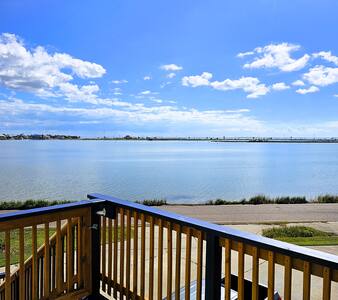 A spacious balcony offers unobstructed views of tranquil waters and a clear blue sky. The railing is made of wooden slats, creating an inviting space for relaxation. The landscape includes grassy areas and a path along the shore, enhancing the outdoor experience.