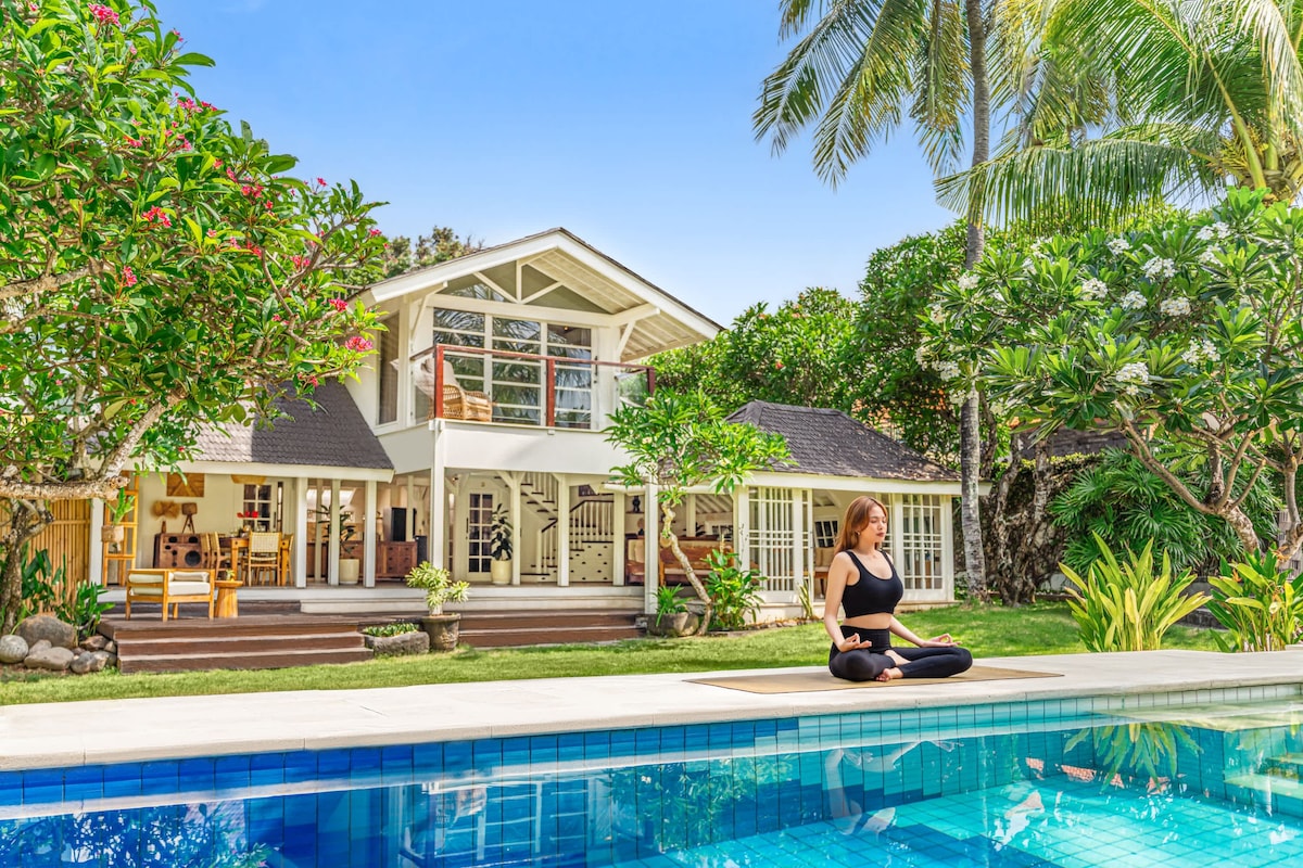 A serene outdoor scene showcases a woman practicing yoga by the pool, surrounded by lush tropical greenery. The two-story villa is visible in the background, featuring large windows and an inviting outdoor dining area. The pool reflects a clear blue sky, enhancing the tranquil atmosphere.
