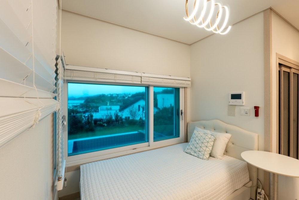 A serene bedroom features a spacious bed with a light quilt and a decorative pillow. Natural light filters through large windows adorned with sheer shades, offering a view of the surroundings. A minimalist nightstand and modern lighting fixture accentuate the calm ambiance.