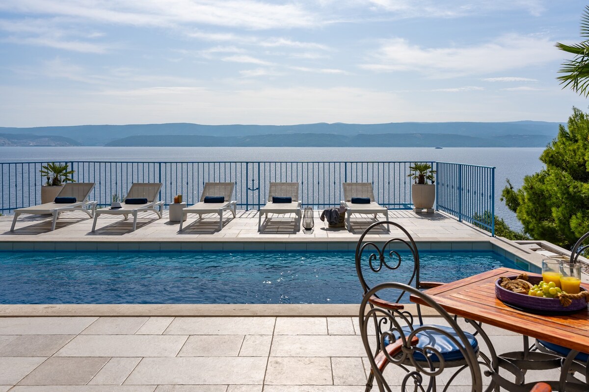 An outdoor area features a private swimming pool surrounded by a spacious deck. Sunbeds are arranged along the deck, offering views of the sea beyond. A dining table set with fresh fruit is positioned nearby, complemented by potted plants and a railing that frames the picturesque landscape.