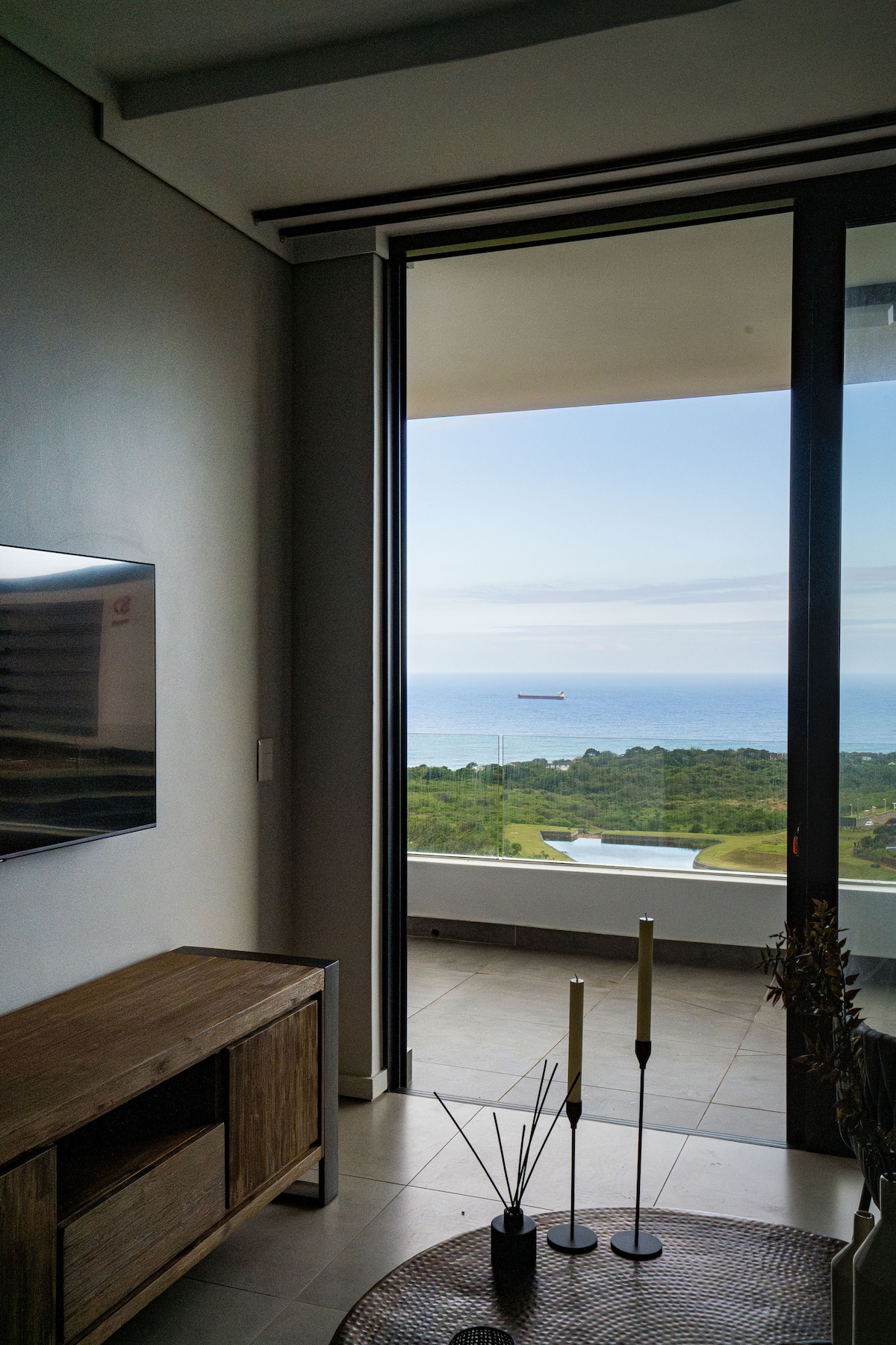 A modern living area showcases floor-to-ceiling glass doors opening to a panoramic ocean view. Natural light brightens the space, complemented by a wooden console table and decorative candles. The serene landscape includes lush greenery and the serene ocean horizon beyond.