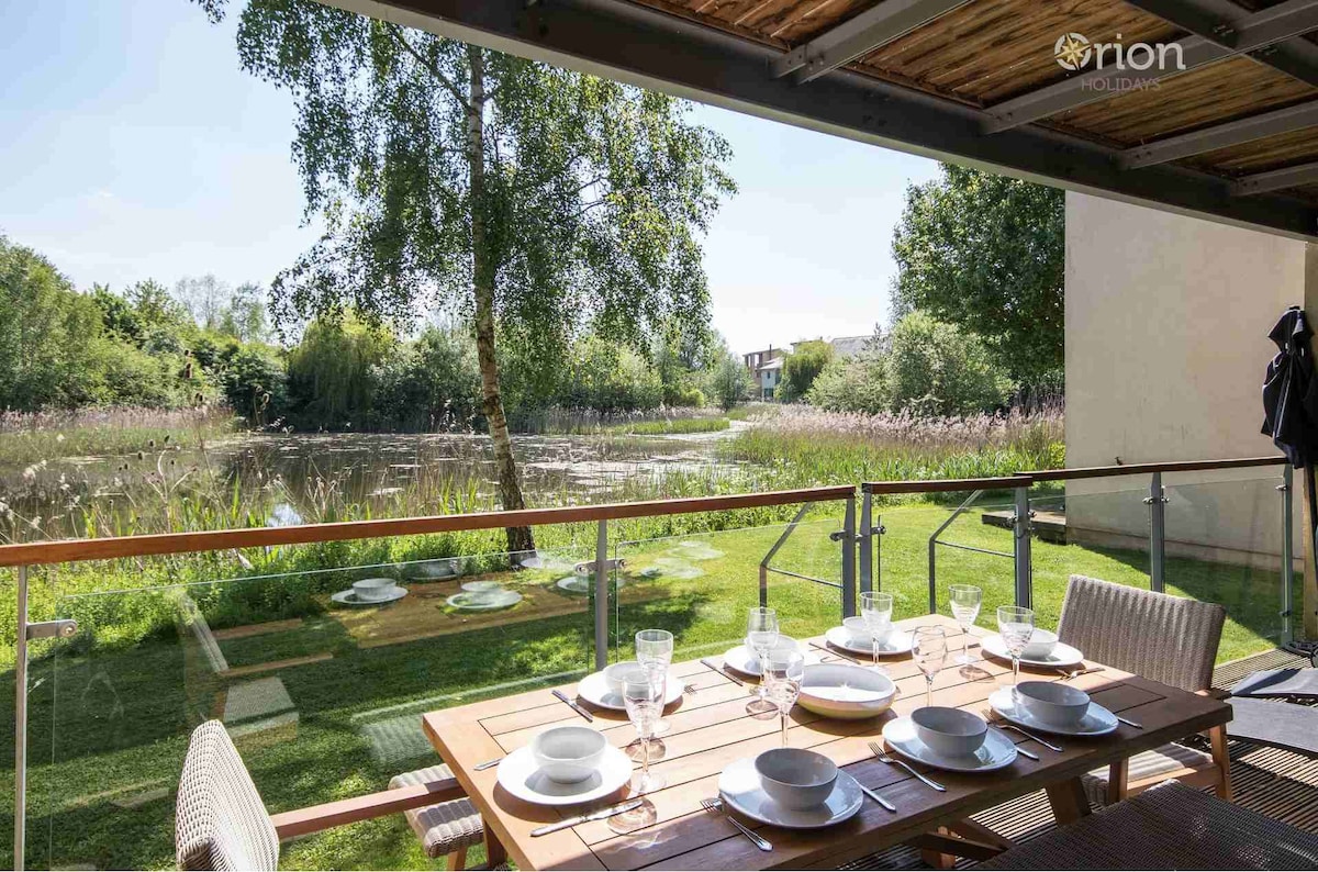 A dining table is set for outdoor meals on a furnished balcony. The scene features a view of the lush grassy area leading to a serene lake, surrounded by trees and natural vegetation. Dishes and glasses await guests to enjoy the tranquil lakeside setting.