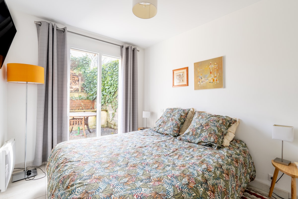 The living/sleeping area features a spacious bed adorned with a patterned duvet. Natural light enters through the sliding glass doors, which lead to a private outdoor terrace, while simple bedside tables are positioned on either side of the bed. Neutral-colored curtains frame the window.